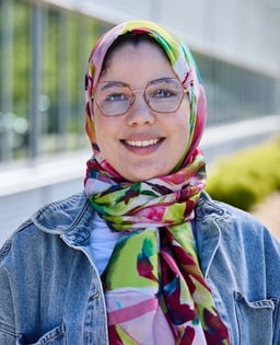 Personne portant un foulard coloré et une veste en jean devant un bâtiment vitré.