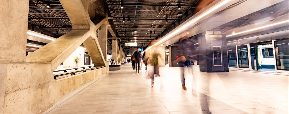 Station de métro moderne avec piliers en béton, passagers en mouvement et train visible.