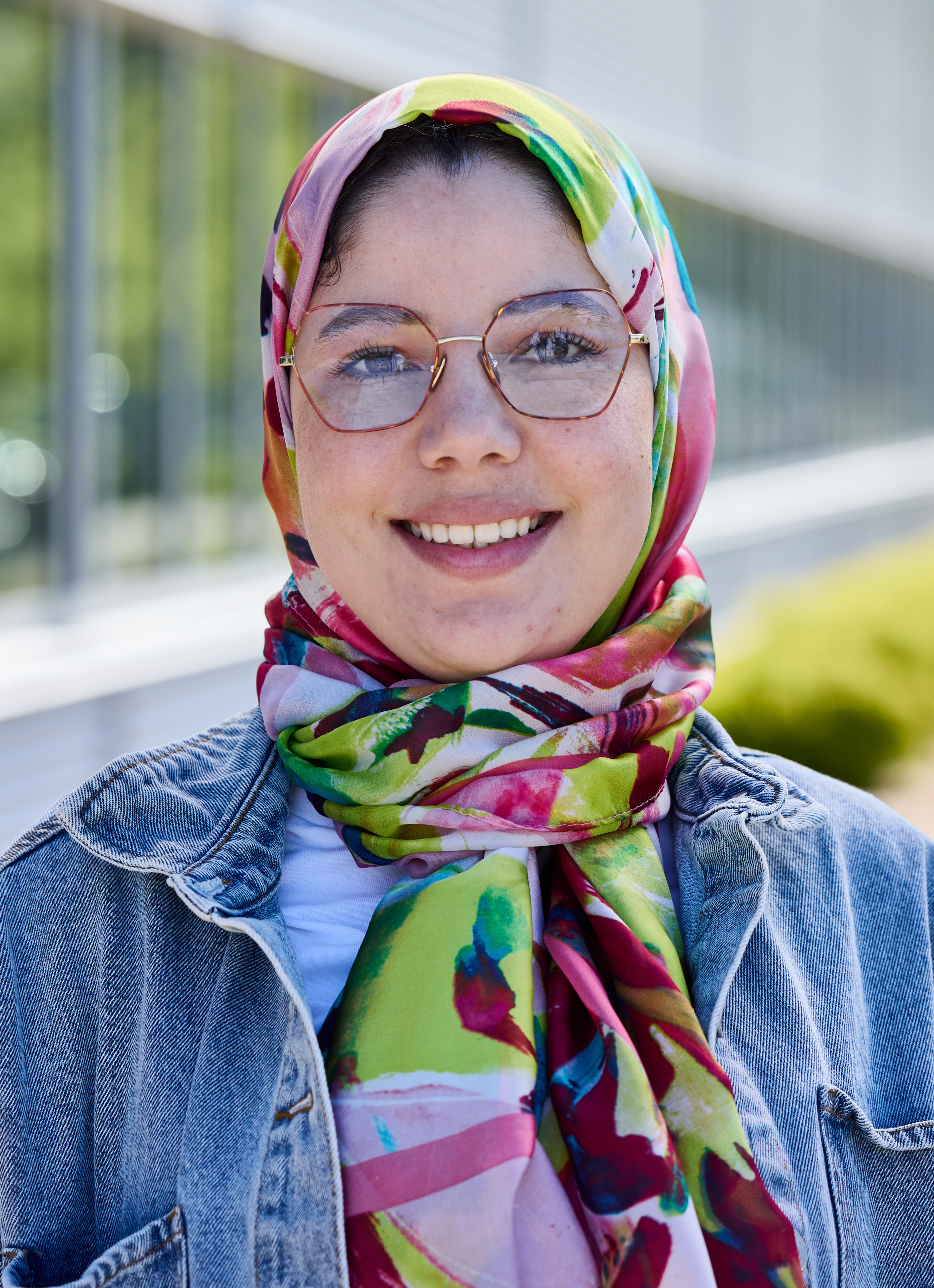 Personne portant un foulard coloré et une veste en jean devant un bâtiment vitré.